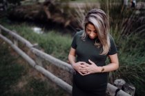 Красивая беременная женщина в повседневном платье улыбается и держит руки на животе, стоя рядом с деревянным мостом — стоковое фото