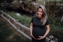 Hermosa mujer embarazada en vestido casual sonriendo y tomándose de la mano en el vientre mientras está de pie junto al puente de madera - foto de stock