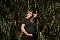 Happy pregnant woman in casual dress stroking belly while standing on green trees wall in sunny summer day with blue sky — Stock Photo