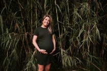 Щаслива вагітна жінка в повсякденному платті погладжує живіт, стоячи на стіні зелених дерев у сонячний літній день з блакитним небом — стокове фото