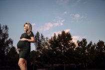 Happy pregnant woman in casual dress stroking belly while standing on pathway in park with green trees — Stock Photo