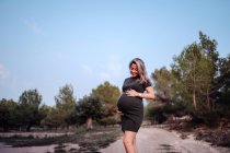 Heureuse femme enceinte en robe décontractée caressant le ventre tout en se tenant sur le chemin dans le parc avec des arbres verts — Photo de stock