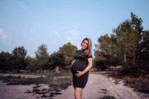 Щаслива вагітна жінка в повсякденному одязі погладжує живіт, стоячи на шляху в парку з зеленими деревами в сонячний літній день — стокове фото
