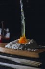 Egg falling over flour while preparing dough on table with eggshell and glass pot on black background — Stock Photo