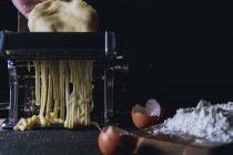 Primer plano de la mano de la persona rodando masa a través de la máquina de pasta mientras se prepara pasta casera fresca en la mesa - foto de stock
