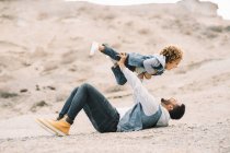Cheerful stylish ethnic man lying down holding curly happy toddler on sandy hills at daytime — Stock Photo