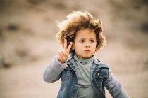 Adorable enfant ethnique frisé occasionnel regardant pensivement loin et faisant signe de paix sur fond flou — Photo de stock