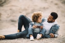 African American casual man leaning on elbow and looking at curly toddler daughter sitting beside with mobile phone — Stock Photo