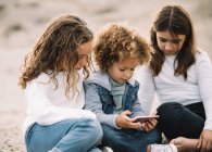 Concentrated casual mixed race kids relaxing on sand and sharing mobile phone at daytime — Stock Photo
