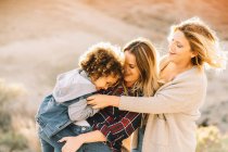 Mãe alegre em camisa quadriculada segurando criança casual como alegre amigo feminino abraçando-os na natureza durante o dia — Fotografia de Stock