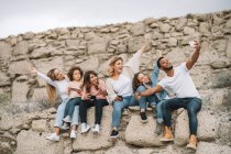 Homme noir prenant selfie avec téléphone portable avec des enfants et des femmes tout en étant assis sur un mur de pierre le jour — Photo de stock