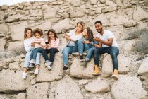 Black man, children and women using gadgets while sitting on stone wall at daytime — Stock Photo