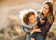 Joyful blonde female holding and cuddling happy child with curly hair looking in camera outdoors at daytime — Stock Photo