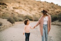 Smiling stylish blonde woman strolling on desert landscape holding hands of surprised toddler with curly hair at daytime — Stock Photo