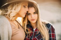 Woman in hat tenderly hugging female with long hair dressed in checkered shirt looking in camera on nature — Stock Photo