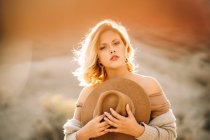 Portrait of graceful female with blonde hair holding hat in nature in backlit sunlight — Stock Photo
