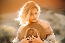 Portrait de femme gracieuse avec des cheveux blonds tenant chapeau dans la nature dans la lumière du soleil rétro-éclairé — Photo de stock