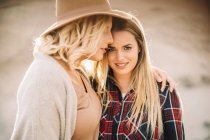 Woman in hat tenderly hugging female with long hair dressed in checkered shirt looking in camera on nature — Stock Photo