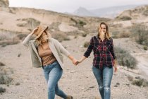 Woman in hat tenderly holding hands with girlfriend dressed in checkered shirt, smiling as walking in nature — Stock Photo