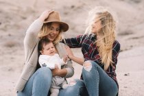Femme heureuse en chapeau assis sur le sable et tenant bébé tandis que l'amie les soutient par temps venteux pendant la journée — Photo de stock