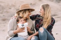 Щаслива жінка в капелюсі сидить на піску і тримає дитину, а подруга-жінка підтримує їх у вітряну погоду вдень — стокове фото