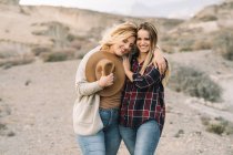 Woman in hat tenderly hugging female with long hair looking in camera dressed in checkered shirt smiling on nature — Stock Photo