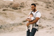 Cheerful bearded African American man in white t-shirt and jeans holding baby in carrier while strolling on nature — Stock Photo