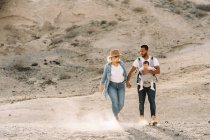 Веселый мужчина, несущий маленького ребенка и держащийся за руки с блондинкой женой во время прогулки по песчаной пустыне — стоковое фото