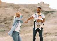 Homem alegre carregando bebê pequeno e de mãos dadas com a esposa loira enquanto dança no deserto arenoso — Fotografia de Stock