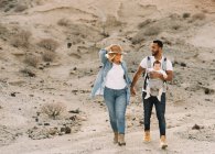 Hombre alegre llevando un bebé y tomándose de la mano con la esposa rubia mientras camina en el desierto de arena - foto de stock