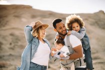 Happy casual Caucasian wife and black smiling husband carrying children on nature at daytime — Stock Photo
