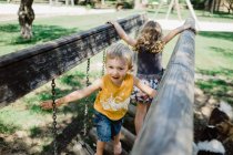 Chico alegre y chica entusiasta escalada jugando y caminando en la cadena de madera en el patio de recreo en el césped verde en el día soleado - foto de stock