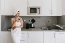 Mujer adulta gozosa disfrutando del día libre mientras toma una bebida caliente después del baño en el apartamento con un diseño interior simple - foto de stock