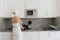 Visão traseira da mulher em toalhas de banho brancas preparando bebidas usando máquina de café em pé na cozinha moderna confortável — Fotografia de Stock
