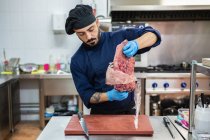 Männlicher Koch in dunkelblauer Uniform und Handschuhen zieht großes Stück rohes Fleisch aus Vakuum-Plastikverpackung — Stockfoto