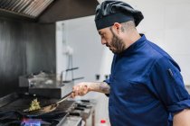 Vista laterale del cuoco maschio in cappello nero scuotendo padella mentre cucina verdure per piatto su pietra a gas in cucina ristorante — Foto stock