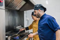 Vista laterale del capo chef maschio che guarda il giovane assistente femminile friggere il cibo mentre si lavora insieme in cucina professionale — Foto stock