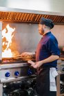 Vue latérale de cuisinier homme faisant flamber sur la nourriture tout en se tenant à la cuisinière à gaz et la préparation de plat dans la cuisine professionnelle — Photo de stock
