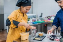 Männlicher Koch beobachtet junge Assistentin mit Löffel in der Hand beim Essen auf Teller legen, während sie gemeinsam in der Restaurantküche arbeitet — Stockfoto