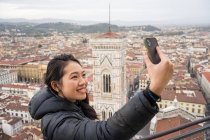 Glückliche asiatische Frau lächelt und macht ein Selfie mit dem Handy, während sie auf der Brunelleschi-Kuppel vor den alten Straßen von Florenz während einer Reise in Italien steht — Stockfoto