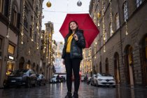 Niedriger Winkel einer glücklichen Frau mit rotem Regenschirm, die lächelt und nach oben schaut, während sie auf der geschmückten Straße in Florenz, Italien, spaziert — Stockfoto