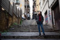 Asian woman with hands in pockets looking away while standing on stairs outside weathered buildings on city street — Stock Photo