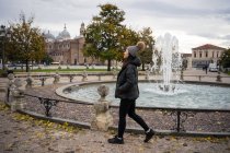 Vue latérale de voyageuse aux cheveux noirs en vêtements chauds et chapeau marchant dans le magnifique vieux parc Prato della Valle à Padova en Italie — Photo de stock
