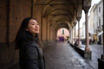Asiatische ruhende Frau lächelt und erkundet alte Straßen mit gerockten Straßen und Gebäuden mit Säulen und schaut weg zu Papua in Italien — Stockfoto