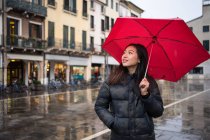 Joven viajera asiática en ropa de abrigo haciendo turismo usando paraguas rojo con edificios antiguos sobre fondo borroso en Padova en Italia - foto de stock