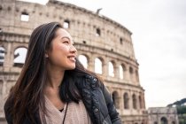 Da soffietto donna felice sorridente e distogliendo lo sguardo mentre in piedi su sfondo sfocato del Colosseo su strada di Roma, Italia — Foto stock