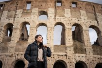 Da soffietto donna felice sorridente e distogliendo lo sguardo mentre in piedi su sfondo sfocato del Colosseo su strada di Roma, Italia — Foto stock