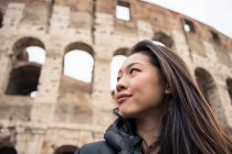 De baixo mulher feliz sorrindo e olhando para longe enquanto estava de pé no fundo borrado do Coliseu na rua de Roma, Itália — Fotografia de Stock