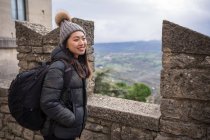 Side view of smiling Asian woman in knitted hat with pompom, warm jacket and backpacking enjoying city in amazing place in San Marino, Italy — Stock Photo