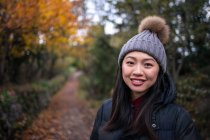 Jovem mulher asiática encantadora em casaco quente preto e chapéu de malha com pompom sorrindo e olhando para a câmera na estrada para o jardim com folhas de outono em San Marino, Itália — Fotografia de Stock